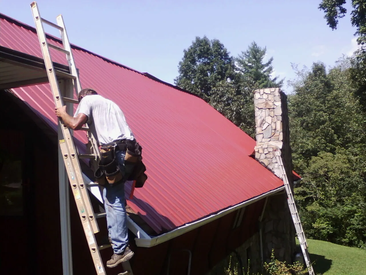 Roofer on a ladder beside a red metal roof and stone chimney — Soffit & Fascia Repair by Roofing Brothers in Sevierville
